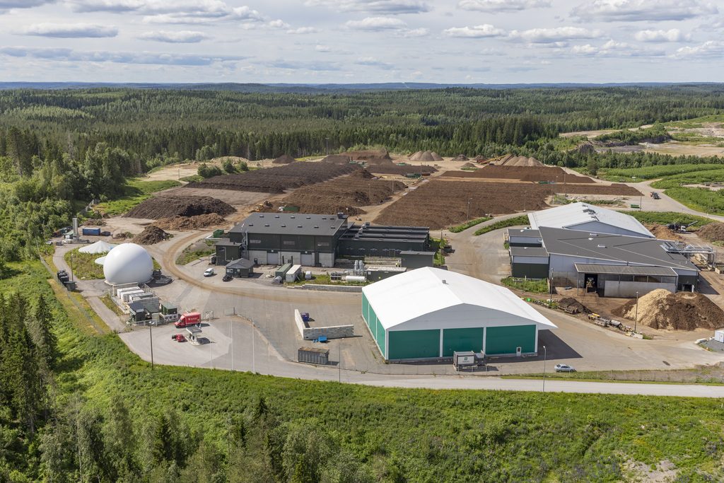Aerial view of a biogas and composting facility surrounded by forest, with production buildings, a gas storage tank, and large compost windrows.
